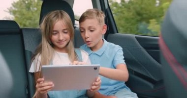 Arka koltukta arabada oturan iki çocuk, tabletlerinin ekranına bakıyorlar. Çocuklar araçları otomatikte kullanıyorlar. Erkek ve kız kardeş birlikte seyahat ediyorlar. Yavaş çekim.