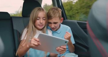 Çocuklar oyun ve eğlence için dijital tablet kullanarak arabada oturur, şarabı kutlar ve arabayla yolculuk ederken beşlik çakar. Aile tatili kavramı. Yavaş çekim.