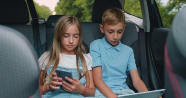 Güzel gülümseyen iki çocuk arka koltukta bir telefon ve tablet kullanarak arabaya biniyor. Küçük çocuk ve kız arabada seyahat ederken telefonda konuşuyor ve bir şeyler gösteriyorlar. Yavaş