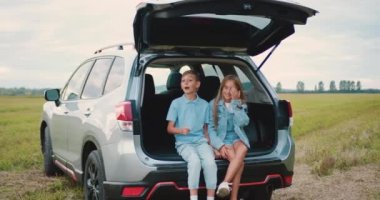 Küçük mutlu çocuklar günlük giysiler giyip yolda arabanın bagajında oturuyorlar ve biraz iletişim kuruyorlar, turistler. Aile gezisi. Yavaş çekim.