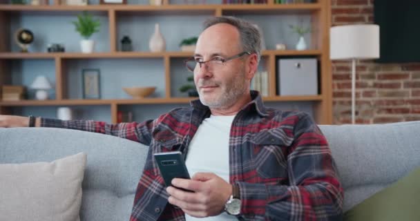 家で快適なソファの上でリラックスした眼鏡で魅力的な満足した現代の大人の髭の男は 夢のようにウィンドウに気を散らし 携帯電話でSmsを書くために瞬間に戻るレジャーの概念 — ストック動画