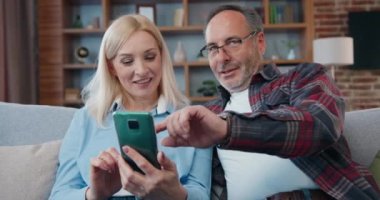 Sevimli, kaygısız, evli bir çift modern oturma odasında rahat bir koltukta oturuyor ve birbirleriyle iletişimin keyfini çıkarıyorlar, telefon uygulamalarında sörf yapıyor, konuşuyor ve gülüyorlar.