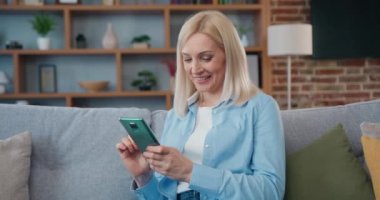 Çekici, açık renk saçlı bir kadının modern oturma odasında yumuşak bir aynasızla dinlendiği ve cep telefonunu arkadaşlarıyla yazışmak için kullandığı ev eğlencesi konsepti.