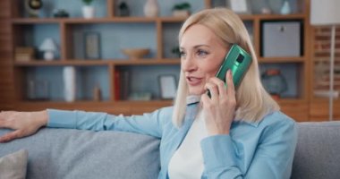 Hoş, gülümseyen, tasasız, açık renk saçlı bir kadının portresi modern dairede rahat bir kanepede rahat rahat sohbet ediyor.