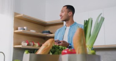 Modern ferah mutfakta duran yakışıklı Afrikalı adam akıllı telefonu tut ve mesaj al. Adam akıllı telefon kullanıyor mutfak tezgahında oturuyor ve evde sağlıklı tarifler arıyor. Yavaş çekim.