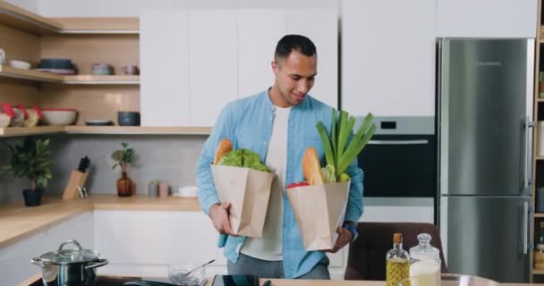 アフリカ系アメリカ人の男が食料品の袋を持って笑顔自宅でモダンなキッチンを歩く — ストック動画