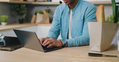 Mutfakta dizüstü bilgisayar ve kulaklık kullanan Afro-Amerikalı bir adam. Gülümseyen Afrikalı siyahi adam evdeki video görüşmesinde dizüstü bilgisayarla buluşuyor. Yavaş çekim.