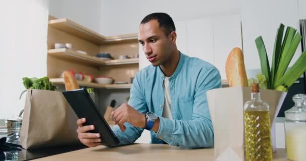 若いハンサムなアフリカ系アメリカ人の男性がモダンなキッチンに座って タブレットコンピュータで良いニュースを読んでいます 感情的な幸せな男は 自宅のオフィスでタブレット上の勝利を祝う スローモーション — ストック動画