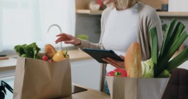 Mutfaktaki tablet bilgisayarla marketten eşyalarını taşıyan ve sebze paketlerini parti çantasına koyan bir kadın. Yavaş çekim.