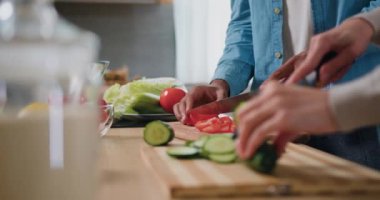 Yakın çekim. Erkek eller domatesleri dilimliyor ve kadın elleri salatalık kesmek için tahta kesme tahtasını salata için kesiyor. Mutfakta sağlıklı yiyecekler var. Yan görüş. Yavaş çekim.