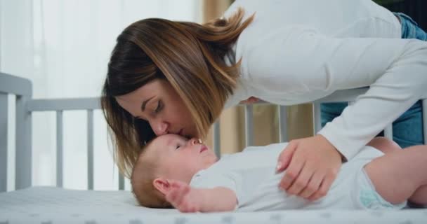 若い母親の近くのUo抱擁し ベビーベッドと笑顔で休んで彼女の愛らしい数ヶ月の赤ちゃんにキス 優しさの概念 — ストック動画