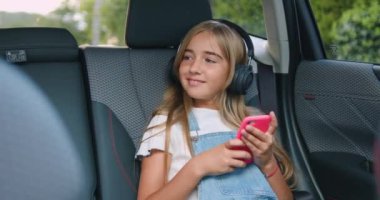 Güzel, tasasız, 12 yaşında, modaya uygun kıyafetleri olan, arka koltukta oturan ve kablosuz kulaklıkla müzik dinleyen, cep telefonunda müzik uygulaması kullanan sarışın bir kız.