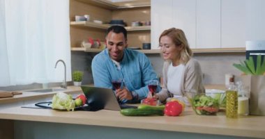 Çok ırklı mutlu çift defter izleyip gülümsüyor ve modern mutfakta şarap içiyor. Genç, sevgi dolu bir aile şarap içiyor, dizüstü bilgisayar kullanıyor, evde yemek yiyor. Yavaş çekim.