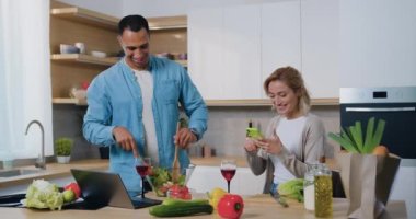 Afro-Amerikalı adam taze salata pişiriyor, sarışın kadın mutfakta oturuyor, şarap içiyor ve akıllı telefon kullanıyor. Evde birlikte gülümseyen genç bir aile. Yavaş çekim.