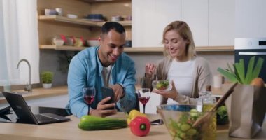 Modern mutfakta telefonu kullanan çok kültürlü genç bir aile. Adam telefonda bir kadına bir şey gösteriyor, kadın gülümsüyor ve salata yiyor. Mutlu aile evde birlikte vakit geçiriyor. Yavaş çekim.