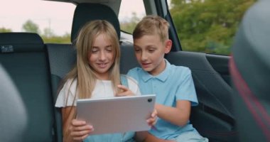 Sevimli, neşeli, kaygısız 10-12 yaşındaki erkek ve kız kardeş arka koltuklarda arabayla seyahat ediyor ve ilginç videolara veya oyun oyunlarına tablet takıyor.