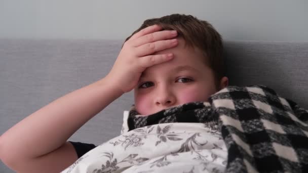 Egy kisfiú beteg, fekszik az ágyban, sálban, tartja a fejét. — Stock videók