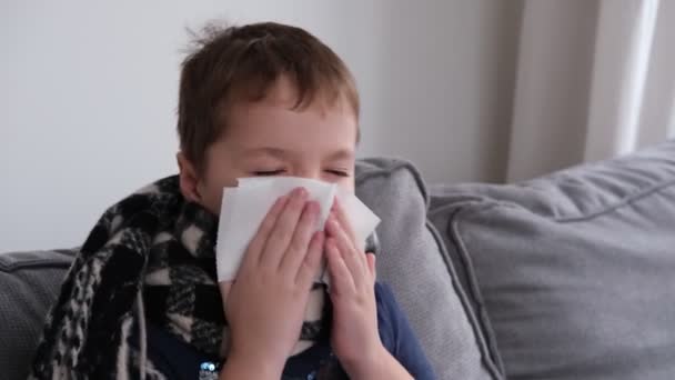 Een kleine jongen is ziek, zit in een sjaal op het bed, blaast zijn neus in een servet — Stockvideo
