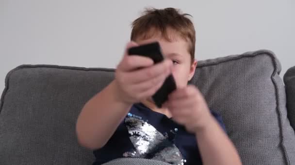 Um garotinho segura um controle remoto nas mãos, troca programas, joga na TV — Vídeo de Stock
