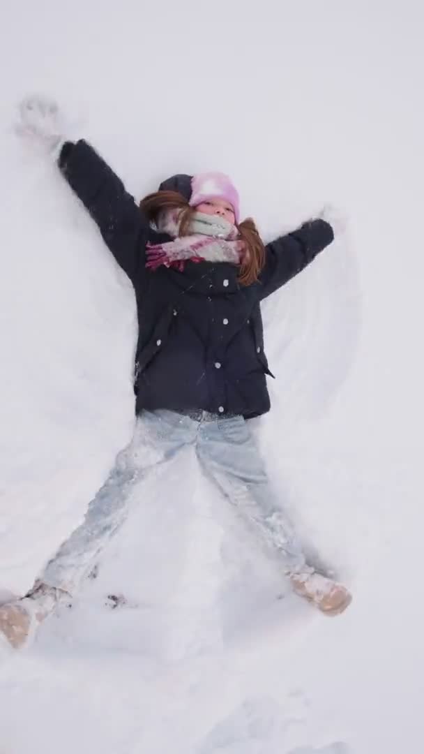 Маленькая счастливая девочка лежит в снегу и делает снежного ангела. Рождество, каникулы, отпуск — стоковое видео