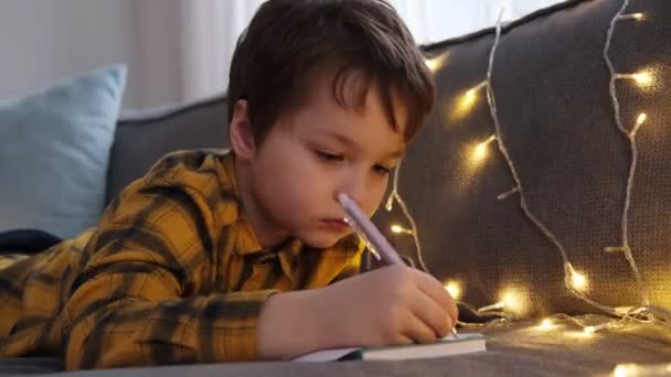 Little boy lies on the sofa and writes a letter to Santa. New Year and Christmas concept — стокове відео