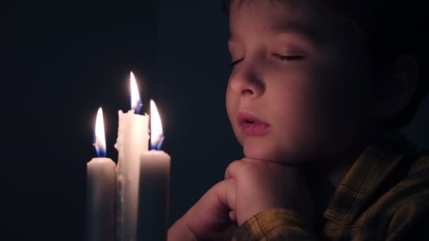 Маленький милий хлопчик читає молитву в темряві зі свічкою — стокове відео