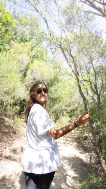 Genç İspanyol kadın gülümsüyor ve ormanda yürüyor. Sonbahar kavramı. Kendi başına güçlü. Beyaz gömlek giyiyor. Dikey. Yüzüne vuran güneş ışığı