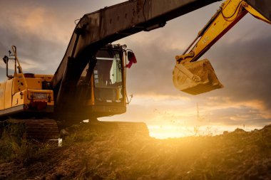 İnşaat alanında kepçeyle toprak kazıyor. Gün batımı gökyüzünde buldozer. Kazıcı inşaat alanına park etmiş. Toprak hareket makinesi. Kovalı kazıcı. Kepçeyle toprak yükleme kovası.