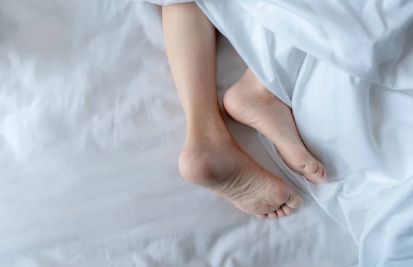 Woman barefoot on bed under white linen blanket in hotel or home bedroom. Healthy sleep and relaxation concept. Lazy Sunday morning. Bare feet of woman chilling sleep on white comfort bed and duvet.