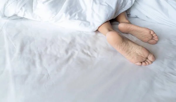 Woman barefoot on bed under white linen blanket in hotel or home bedroom. Healthy sleep and relaxation concept. Lazy Sunday morning. Bare feet of woman chilling sleep on white comfort bed and duvet.