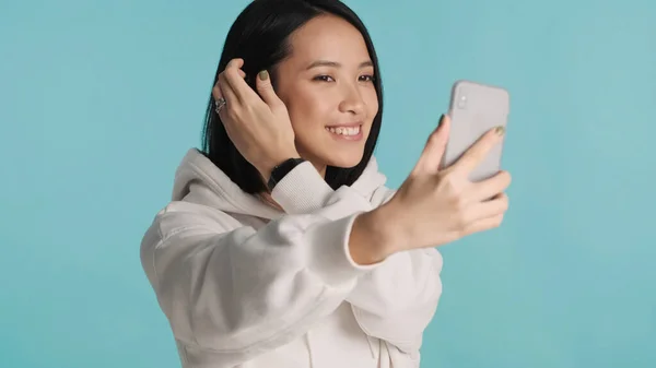 Asian female wearing hoodie preening and taking selfie on smartphone over colorful background. Modern technology concept. Beautiful woman posing in studio