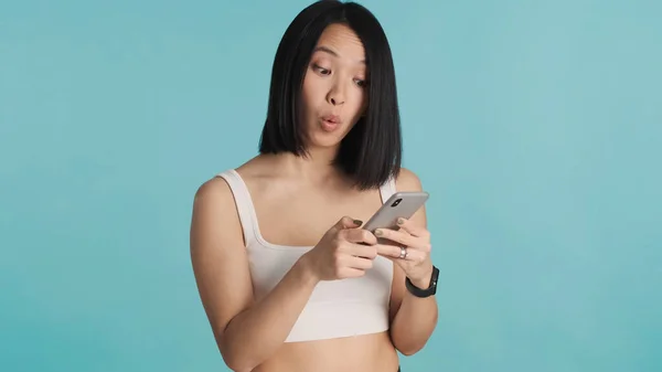 Surprised Asian woman staring at smartphone reading news over blue background. Wow face expression