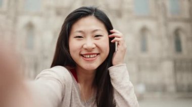 Esmer Asyalı kız, eski mimarinin arka planında selfie çekerken samimi bir şekilde gülümsüyor. Japon kadın yeni şehri keşfediyor.