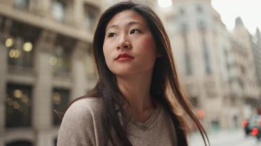 Düşünceli Asyalı uzun saçlı kadın arka planda geçen arabalarla caddede dikiliyor.