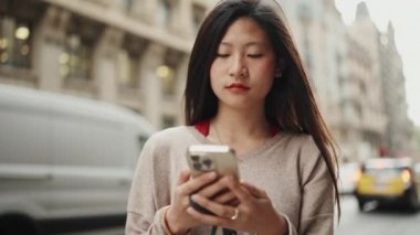 Uzun saçlı güzel bir Asyalı kadın arka planda geçen arabalarla sokakta akıllı telefon kullanarak sosyal medyasını kontrol ediyor. Asyalı kız dışarıda taksi bekliyor.