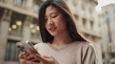 Uzun saçlı güzel bir Asyalı kız sokakta akıllı telefon kullanarak en iyi arkadaşıyla mesajlaşıyor. Genç Asyalı kadın gülümsüyor ve dışarıdaki kameraya bakıyor.