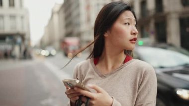 Güzel, uzun saçlı Asyalı kadın akıllı telefondan haritayla arka planda geçen arabalarla caddede doğru yeri arıyor.