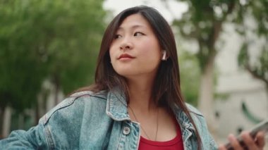 Kablosuz kulaklık takan uzun saçlı Asyalı kadın bankta oturmuş iyi müzik dinliyor. Kaygısız kız dışarıda dinleniyor..