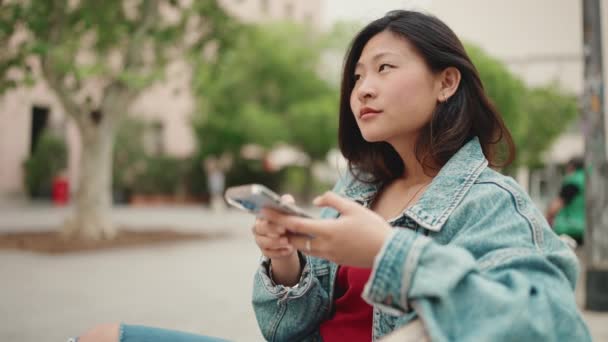 Długowłosy Azjatka Kobieta Dżinsowej Kurtce Siedzi Ławce Zewnątrz Sprawdzając Swój — Wideo stockowe