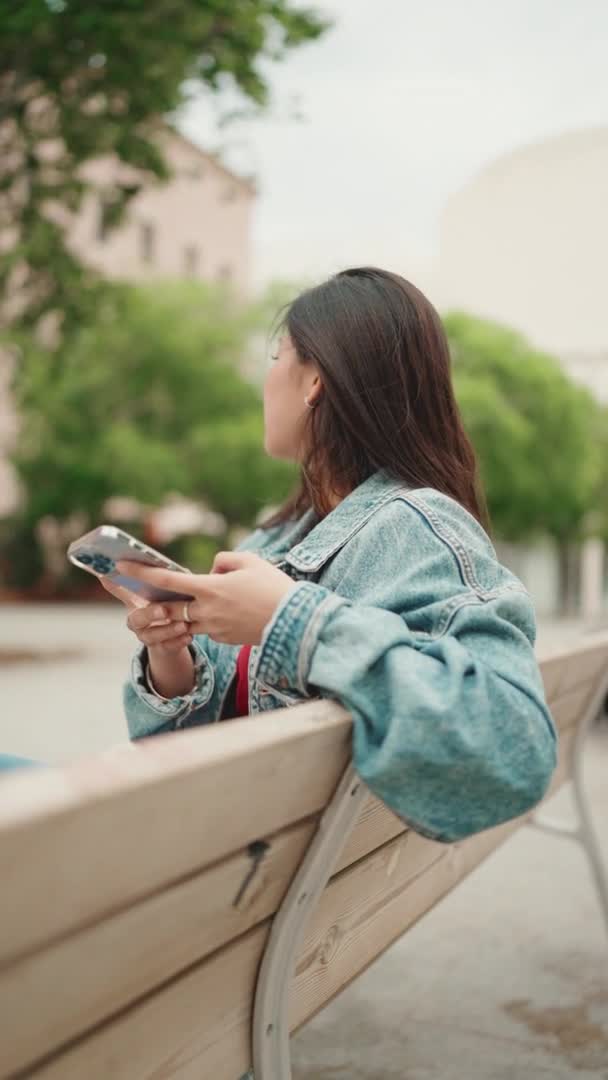 Pionowe Strzał Długowłosy Azji Kobieta Dżinsowej Kurtce Siedzi Ławce Smartfonem — Wideo stockowe