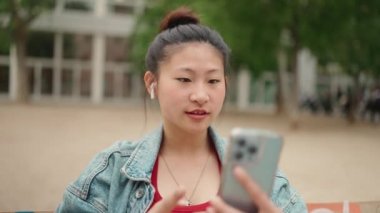 Kablosuz kulaklık takan güzel Asyalı kız dışarıda akıllı telefon kullanarak sosyal medyası için öpüşme videosu gönderiyor. Kadın blog yazarı yeni içerik yaratıyor