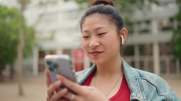 Wesoła Azjatka Słuchawkach Siedząca Zewnątrz Używająca Smartfona Koncepcja Nowoczesnej Technologii — Wideo stockowe