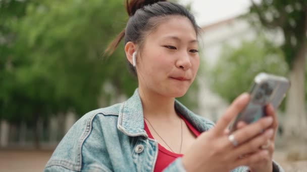 美しいアジアの女性は スマートフォンをチェックし 屋外で笑顔の無線イヤフォンを身に着けている 近代技術の概念 — ストック動画