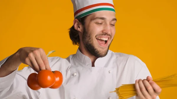Close up Italian chef wearing uniform looking excited holding tomatoes and pasta in hands over colorful background