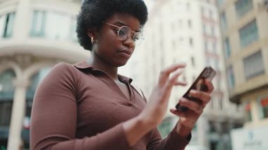 Sokakta akıllı telefonuyla duran gözlüklü çekici Afrikalı Amerikalı kız. Açık havada telefon kullanan esmer tenli kadın.