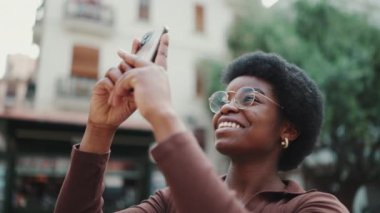 Afro-Amerikalı kıvırcık kız telefonda güzel bir mimarinin fotoğrafını çekerken heyecanlı görünüyor. Sokakta gülümseyen gözlüklü güzel kadın.