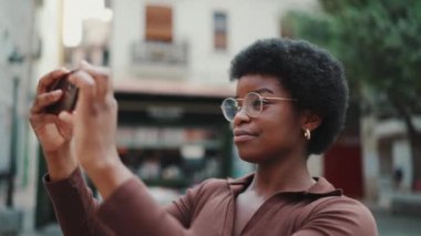Sokakta yürürken gözlüklü Afro-Amerikan kıvırcık kızın fotoğrafını çekiyorum. Dışarıda akıllı telefon kullanarak gülümseyen genç bir kadın.