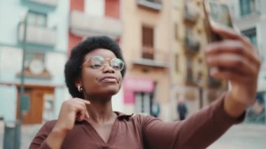 Sokakta selfie çeken Afro-Amerikan kıvırcık kadın portresi. Güzel kadın blogcu dışarıda fotoğraf çekerken barış jesti gösteriyor