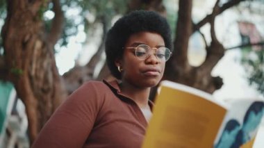 Sokakta kitap okuyan güzel bir Afrikalı kadının portresi. Koyu tenli, gözlüklü, açık havada okuyan kız öğrenciyi kapat.