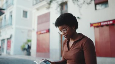 Gözlüklü güzel Afrikalı Amerikalı kadın sokakta oturmuş kitap okuyor. Açık havada okuyan esmer tenli bir kız.